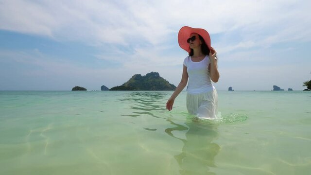Tourist woman wearing white clothes slowly walk at waist deep water, along sea beach. She hide from hot tropical sun under wide hat. Crystal clear waters, islands on background