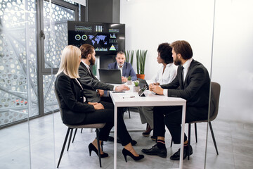 Team of diverse five multiethnic businesspeople having meeting in boardroom at office indoor, sitting at the desk with tablets and laptop, in front of a huge plasma TV screen