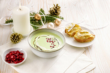 Fototapeta na wymiar Cream soup from apples and peas with pomegranate seeds and baked camembert canapes, appetizer for a holiday meal on a white wooden table with burning candle and Christmas decoration, copy space