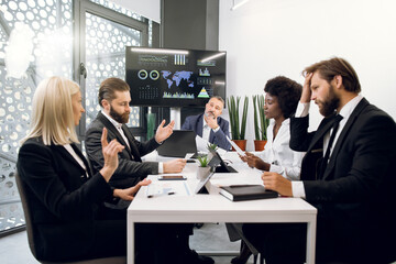 Confident male and female multiethnic economists or directors, discussing company strategy during brainstorming at office, with world map and statistic charts on wall TV screen on the background
