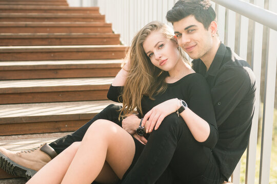 Beautiful young multiracial couple, student couple in love, sit wooden staircase in the city. Beautiful Turkish brunette guy hug a Caucasian girl in black clothing. Date young people in sunny weather