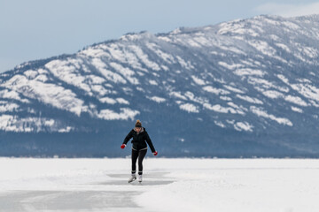 Woman skating on frozen lake in northern Canada with huge mountains in the background surrounded by snow, snow capped and glassy ice on lakes surface. 