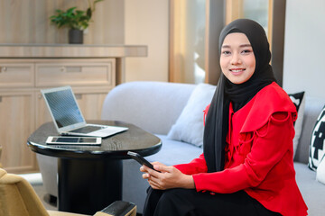 Portrait of beautiful young business woman in red hijab holding smartphone at living room, messaging or working with networks, smiling Muslim female sitting on sofa in lobby hotel