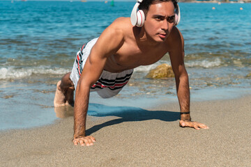 Fototapeta na wymiar Sporty Latin man in headphones doing push-ups on seashore