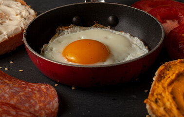 close up macro image of a fried egg (sunny side up) in a non stick pan on black wooden table. tomatoes, buttered and creamed toasts and slices of salami are seen in the background