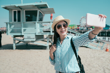 happy young female travel backpacker hold cellphone taking selfie with open lifeguard station....