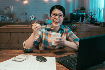 hungry businesswoman in glasses eating instant noodles fast food in disposable cup while working and using computer laptop and smiling face camera. happy young asian chinese girl holding ramen soup