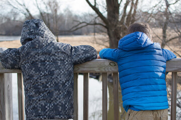 Two boys at a nature preserve looking over a railing at a pond and trees in winter © Julia Beatty