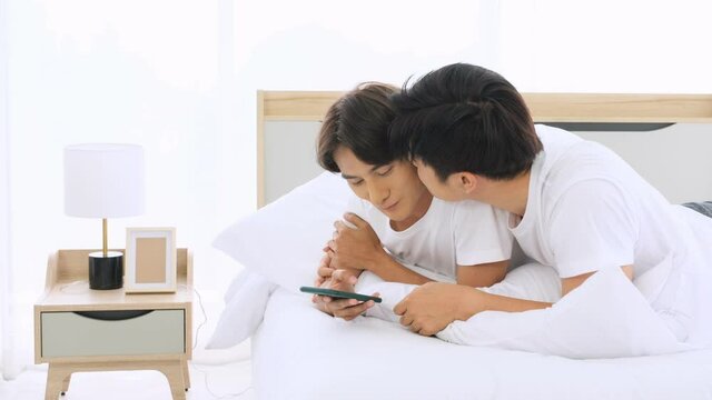 Gay couples use phone and lying down happily chatting on bed in the morning.