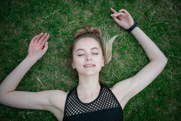 Happy beautiful young teen girl is lying on the green grass in summer with her hands up and resting. The view from the top.