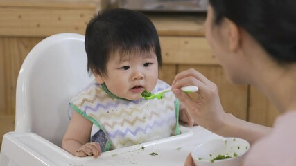 cropped shot asian new mother is guiding and encouraging her baby to eat while it is looking at the solid food with hesitation on the baby chair.
