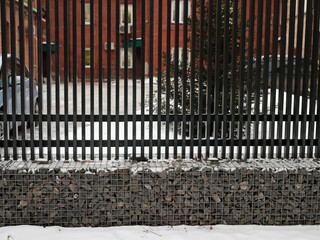 Fence with a gabion structure. Materials - metal, stone. Background - a building, a green fir-tree, cars. White snow in winter.