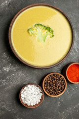 Vertical view of creamy broccoli soup in a brown bowl and different spices on gray table