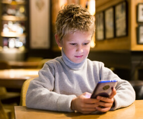 Portrait of tween boy carried away with phone while sitting at table in cafe..