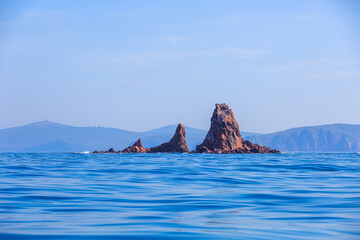A picturesque red stone island sticks out of the blue sea water. An uninhabited rocky island in the middle of the blue sea against the backdrop of a clear sky.