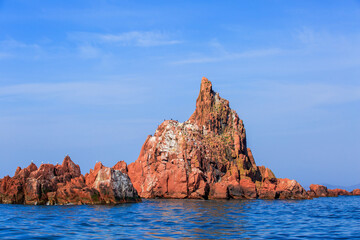 A picturesque red stone island sticks out of the blue sea water. An uninhabited rocky island in the middle of the blue sea against the backdrop of a clear sky.