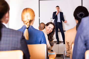 Portrait of cheerful young woman peeking from front row in crowded conference hall during lecturer...