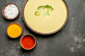 Overhead view of creamy broccoli soup in a brown bowl and different spices on gray table