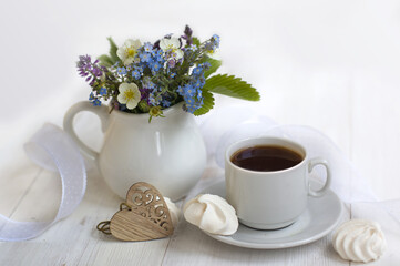 Obraz na płótnie Canvas romantic breakfast: cup of coffee, meringues, a jug with forget-me-nots, wild strawberry flowers, white ribbon, carved wooden heart on a light background