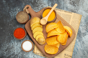 Above view of tasty homemade chips cut potato slices on wooden cutting board and different spices on newspaper on gray background