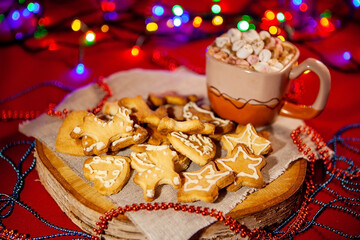 Christmas gingerbread cookies on a wooden stand and a cup of hot chocolate with marshmallows in the back, on a red background with a beautiful bokeh of glowing garland. Selective focus. Christmas and 