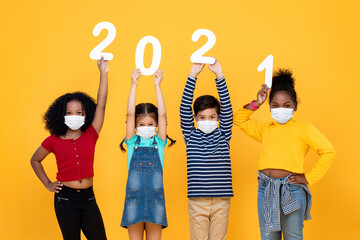 Cute mixed race children wearing medical face masks and holding 2021 numbers for corona virus pandemic protection in new year celebration concept isolated on yellow background