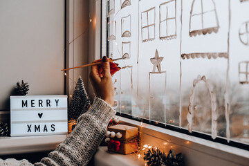 Female hand draws christmas holidays decoration on window glass. New Years scenery. city buildings...