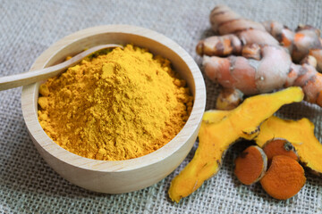 Close up the turmeric dry powder in wooden bowl and fresh turmeric and cutting of turmeric roots on the sackcloth background. selected focus.