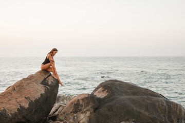 Fototapeta na wymiar Beautiful woman in black swimsuit posing on the beach sitting on rocks, ocean view landscape
