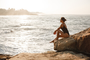 Beautiful woman in black swimsuit posing on the beach sitting on rocks, ocean view landscape