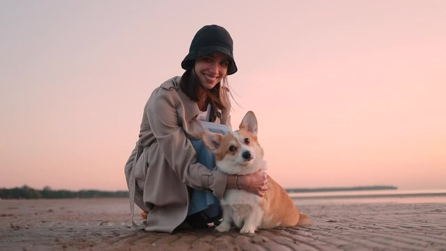 Corgi dog and woman in hat posing on beach at sunset spbi. Portrait of couple who spend time in nature in fall. Pet and beautiful happy female sitting on sand by sea. Caucasian lady in raincoat