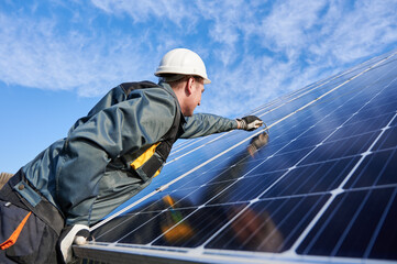 Side view snapshot of workman, wearing uniform, working gloves and helmet, setting a shiny new solar battery with help of hex key, blue sky on background. Green energy concept