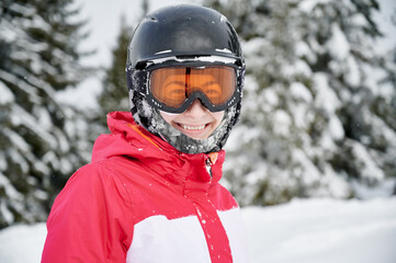 Fototapeta na wymiar Portrait of smiling girl wearing red jacket, helmet and goggles in snowy mountains, close-up. Concept of winter kinds of sport