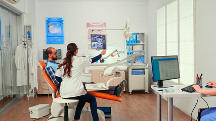 Doctor comming in consultation room examining patient with dental bib before dental intervention sitting on stomatological chair while stomatologist nurse making appointments writing on computer.