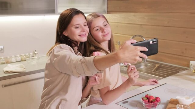 Medium shot of enjoying mom and daughter taking selfie while eating delicious strawberry popsicle in home kitchen