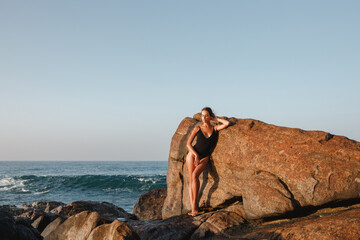 Woman model in black  swimsuit is posing on the beach with big stones in background. Beautiful tanned girl. Concept bikini, swimwear