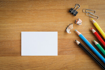 Table top view, on a wooden desk lay down multi colored pencils, clips and a white card, table top...