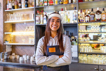 Portrait of a beautiful bartender standing at the counter smiling and looking at the camera while while wearing face shield due to covid-19, shelves full of bottles with alcohol on the background