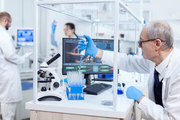 Experienced scientist working in busy laboratory at his workplace holding dropper dispense. Senior professional chemist using pippete with blue solution for microbiology tests.