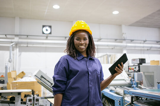 Happy black female factory worker in hardhat and overall, standing at machine, holding tablet, looking at camera and smiling. Front view, medium shot. Women in industry concept