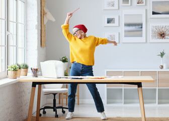 cheerful elderly woman freelancer creative designer in a red hat having fun and dancing in...