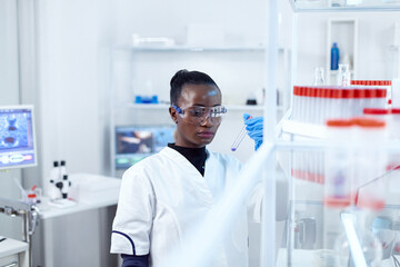 African scientist holding test tube with blue liquid for medical purpose. Black researcher in sterile laboratory conducting pharmacology experiment wearing coat.