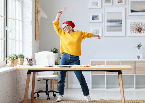 cheerful elderly woman freelancer creative designer in a red hat having fun and dancing in workplace.