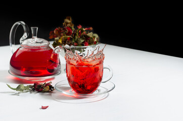 Autumn evening teatime with splash in red herbal tea cup, transparent tea pot with hawthorn berries, rose hip, dry leaves on white wood table, black wall.