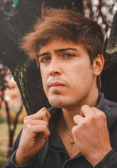 Serious guy posing staring in camera with hands on shirt lapel in a park.