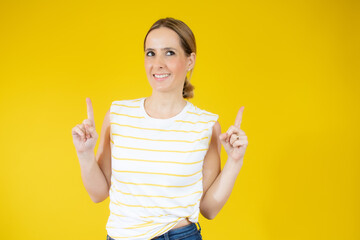 Portrait of a happy young woman dressed in casual t-shirt pointing fingers up at copy space isolated over yellow background