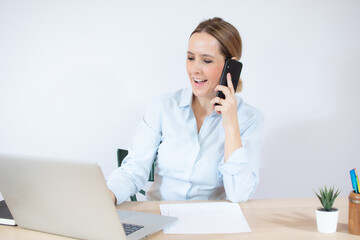 Good business talk. Cheerful young beautiful woman talking on mobile phone smiling and using laptop while sitting at her working place