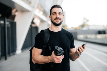 Fototapeta premium Cheerful man looking at the camera and smiling. Young male athlete in black clothes holding sports bottle and smartphone in hands. Attractive man is waiting for a friend to start morning run together.