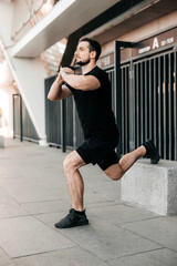 Athletic man stretching legs before workout outdoors. Active living. Urban sport concept. Runner in black sportswear excercising at morning holding hands in lock. Morning warm up.