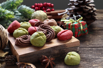 homemade colorful chocolate, green tea and coffee cookies are on the small wooden slats with chritmas ornaments decoration , cinnamon, pinecone, star anise and chritmas ornament on the wooden table